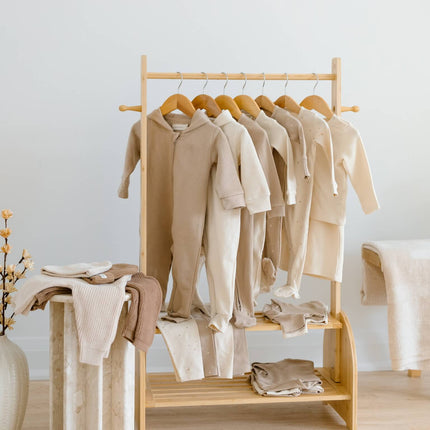 A light natural wood clothing rack holding multiple cream and beige organic cottan baby garments, including long-sleeve tops and folded pieces on the lower shelf, in a softly lit minimalist room. there are 23 pieces in this kindnest capsule