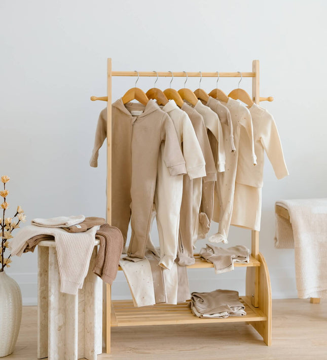 A light natural wood clothing rack holding multiple cream and beige organic cottan baby garments, including long-sleeve tops and folded pieces on the lower shelf, in a softly lit minimalist room. there are 23 pieces in this kindnest capsule