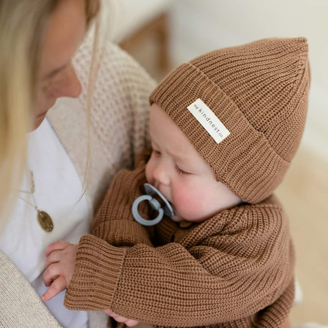 A baby wearing a brown organic cotton ribbed knit beanie with a white 'THE KINDNEST CO.' label and a matching organic cotton brown knit cardigan, held by a woman in a cream knit cardigan.