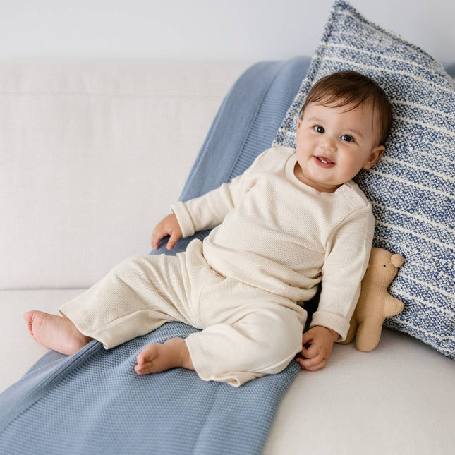 A baby wearing a cream-colored long-sleeve organic cotton waffle knit shirt and wide leg waffle knit pants on a white sofa, leaning against a blue and white striped pillow and a blue knit blanket.