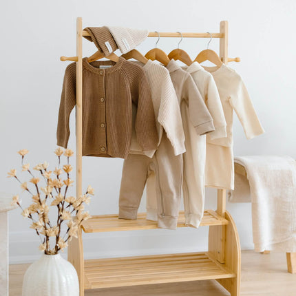 A light natural wood clothing rack holding several organic cotton baby garments in neutral tones, including a brown cardigan, cream and beige tops, and pants, set in a bright room with a white vase of dried flowers and a white chair in the background. There are 7 pieces in total, in this Kindnest capsule