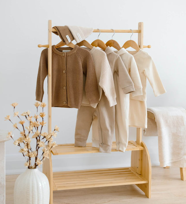 A light natural wood clothing rack holding several organic cotton baby garments in neutral tones, including a brown cardigan, cream and beige tops, and pants, set in a bright room with a white vase of dried flowers and a white chair in the background. There are 7 pieces in total, in this Kindnest capsule