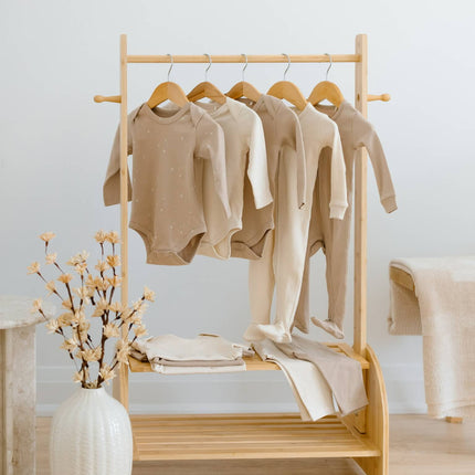 A light natural wood clothing rack with shelves and wooden hangers, holding several beige and cream organic cotton baby garments. The rack is styled in a bright, minimalist room with a white vase of dried flowers on a light wood floor. This Kindnest Capsule has 10 items in total. 