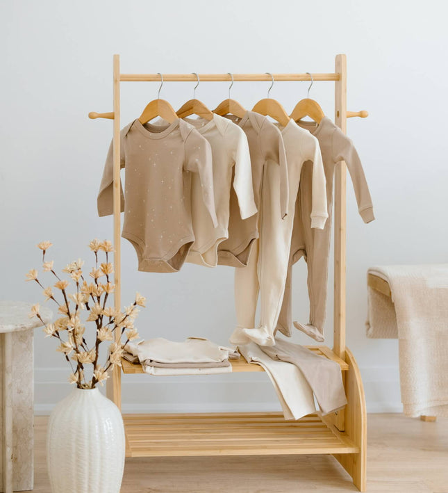A light natural wood clothing rack with shelves and wooden hangers, holding several beige and cream organic cotton baby garments. The rack is styled in a bright, minimalist room with a white vase of dried flowers on a light wood floor. This Kindnest Capsule has 10 items in total. 