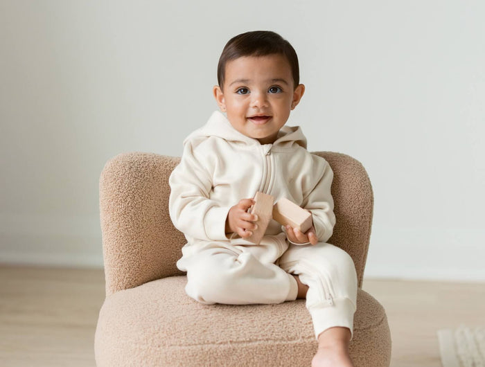 A baby sitting on a small beige boucle armchair in a minimalist room, wearing The NOA Romper by The Kindnest Co, an organic baby clothign brand.