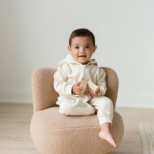 A baby sitting on a small beige boucle armchair in a minimalist room, wearing The NOA Romper by The Kindnest Co, an organic baby clothign brand.