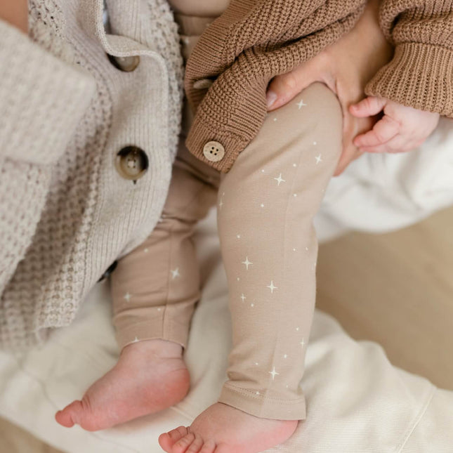 A close-up lifestyle shot of a baby wearing beige organic cotton leggings with tiny white star prints, styled with a brown organic cotton chunky knit cardigan.
